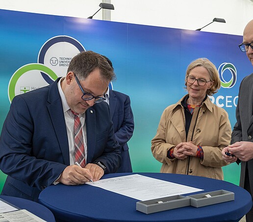Rektor Kratzsch unterschreibt ein Dokument auf einem blauen Stehtisch umringt von anderen Beteiligten des Projekts.
