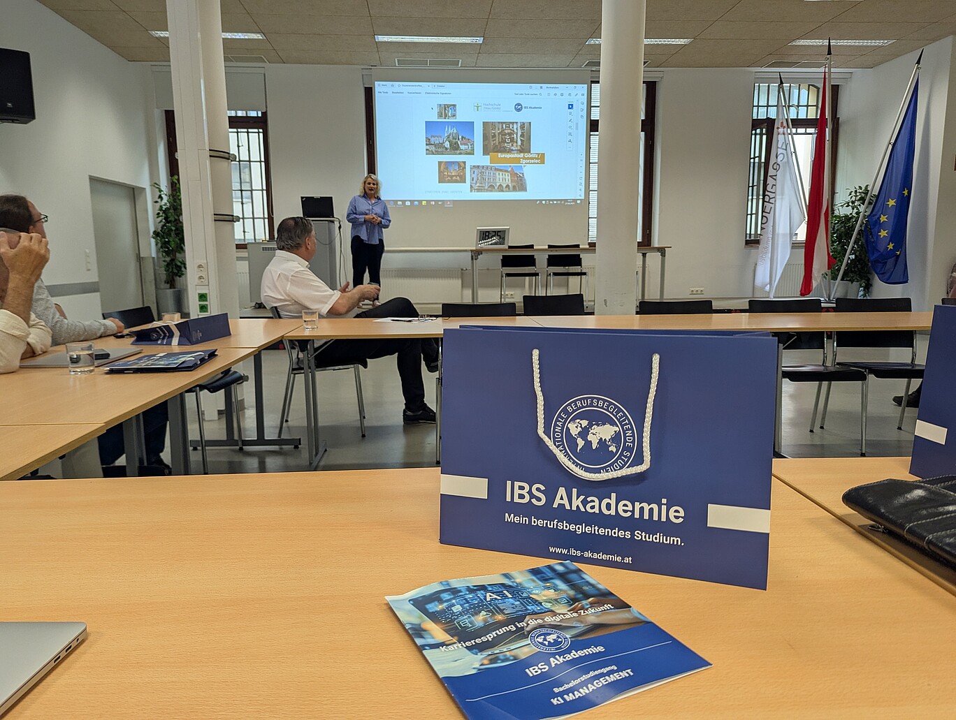 Seminarraum mit Teilnehmenden, die einer Präsentation folgen. Im Vordergrund eine blaue Geschenktasche mit dem Logo der IBS Akademie und der Aufschrift „Mein berufsbegleitendes Studium“. Im Hintergrund steht eine Referentin vor einer Projektionsfläche mit Präsentationsfolien. Auf dem Tisch liegen Info-Broschüren und Materialien zum Studiengang „KI Management“.