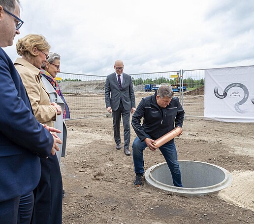 Ein Mann steht mit einem Bein in einem aus der Erde ausgehobenen Loch und hält den Grundstein in den Händen. Drumherum stehen die Beteiligten.