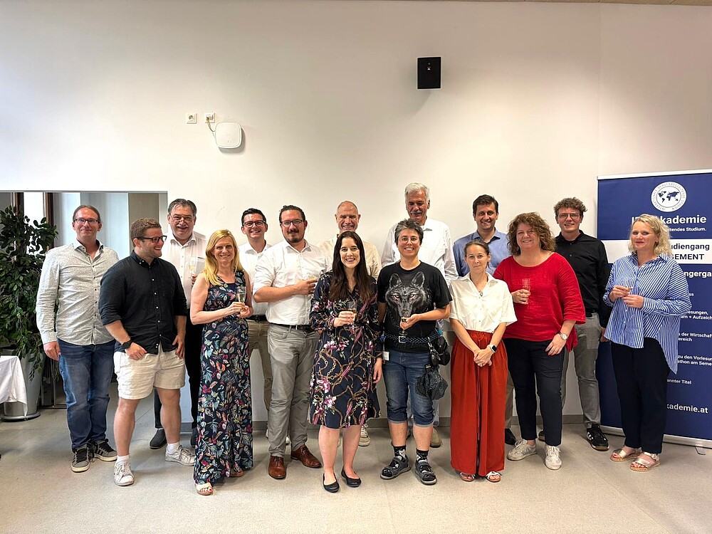 Gruppenfoto von 17 Personen bei einer Veranstaltung in einem Seminarraum. Die Personen stehen lächelnd beisammen, einige halten ein Getränk in der Hand. Im Hintergrund ist ein Roll-Up mit dem Logo der „FH Akademie“ zu sehen, was auf einen akademischen oder beruflichen Anlass hinweist, z. B. Abschluss oder Netzwerktreffen.