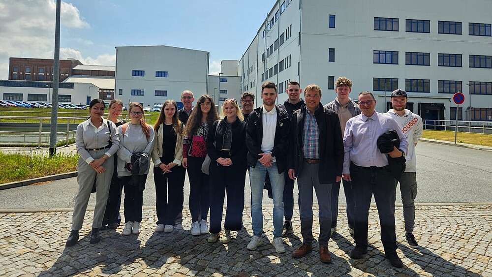 Eine Gruppe Studierender und Lehrender stehen auf einem Platz vor den Produktionsgebäuden der fit GmbH.