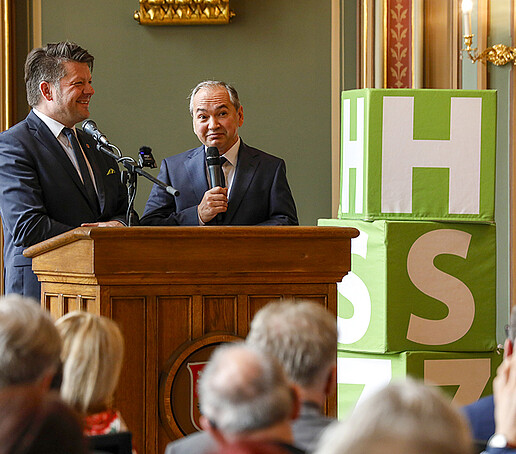 Herr Zenker und Herr Ursu stehen zusammen am Rednerpult und sprechen zum Publikum.