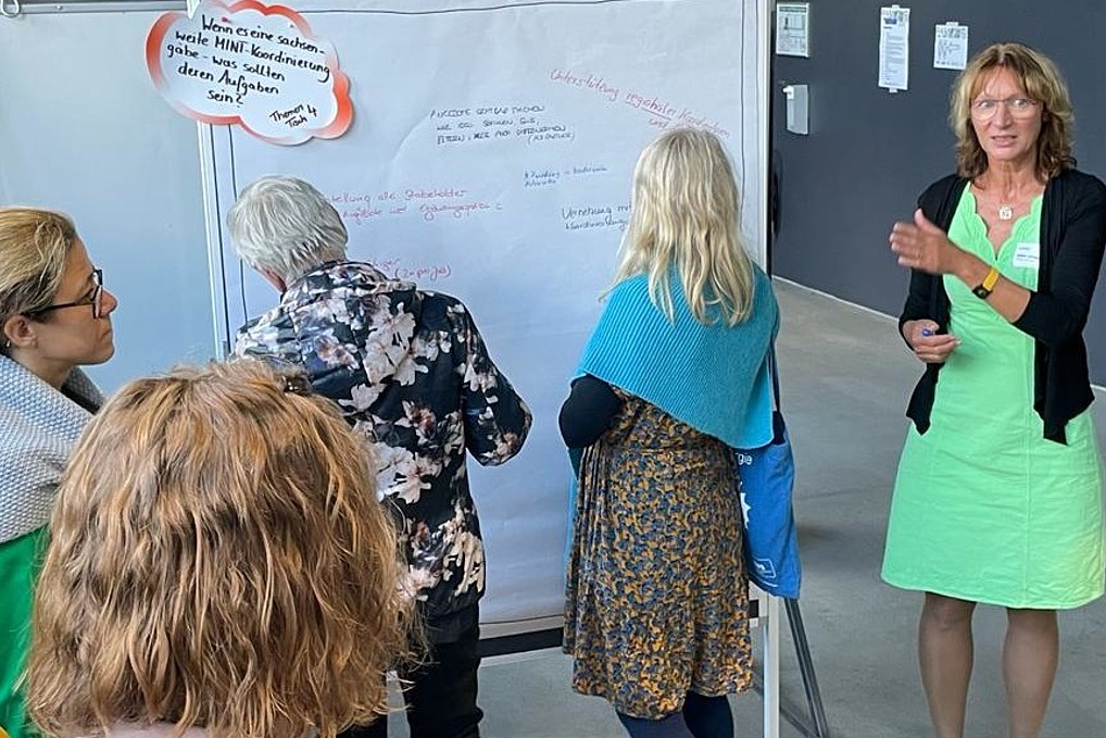 Sabine Scholz und Workshop-Teilnehmende vor einem Whiteboard, auf das gerade eine Frau etwas schreibt.