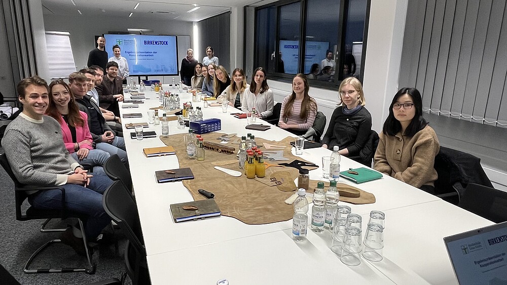 Das Bild zeigt eine Gruppe von jungen Erwachsenen, die um einen Konferenztisch sitzen, auf dem verschiedene Materialien und Getränke stehen, während zwei Personen an einer Präsentationswand stehen.  Im Detail zeigt das Bild eine große Gruppe von etwa 20 jungen Erwachsenen, hauptsächlich Frauen, die um einen langen, weißen Konferenztisch sitzen. Die meisten Teilnehmer sind in legerer, aber gepflegter Kleidung gekleidet. Der Tisch ist mit verschiedenen Gegenständen bedeckt: mehrere Notizbücher, Wasserflaschen, kleine Flaschen mit Getränken (vielleicht Säfte oder Liköre), Gläser und eine große braune, offenbar aus Leder bestehende Fläche, die wahrscheinlich ein Präsentationselement ist und verschiedene Lederstücke und Materialien zeigt – möglicherweise im Zusammenhang mit dem auf der Präsentationswand sichtbaren Logo von Birkenstock. Die Atmosphäre wirkt entspannt und kollegial, eher ein informelles Treffen als eine strenge Konferenz. Im Hintergrund stehen zwei Personen vor einer Präsentationswand, auf der das Logo von Birkenstock und die Worte „Ergebnissepräsentation der Kooperationarbeit“ zu sehen sind, was auf ein Treffen im Zusammenhang mit einem Projekt oder einer Zusammenarbeit schließen lässt. Die Beleuchtung ist hell und neutral. Die Gesamtstimmung ist positiv und produktiv.