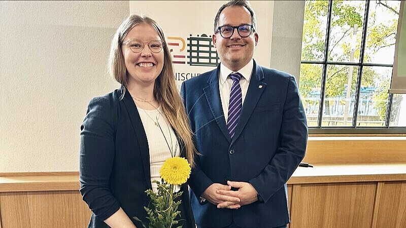 Professorin Schellhammer steht neben Rektor Kratzsch und hält eine gelbe Blume in der Hand, beide lächeln in die Kamera.