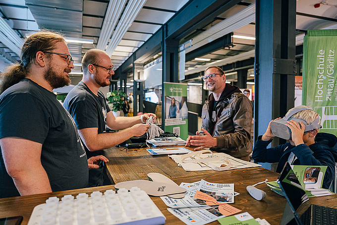 Drei Menschen unterhalten sich am Stand des AAL-Labor. Ein Kind probiert die VR_Brille aus