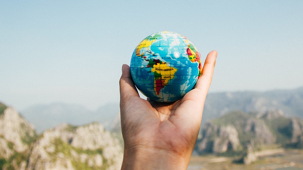 Eine ausgestreckte Hand hält einen Erdglobus, im Hintergrund sieht mein steinige Gebirgsformationen.