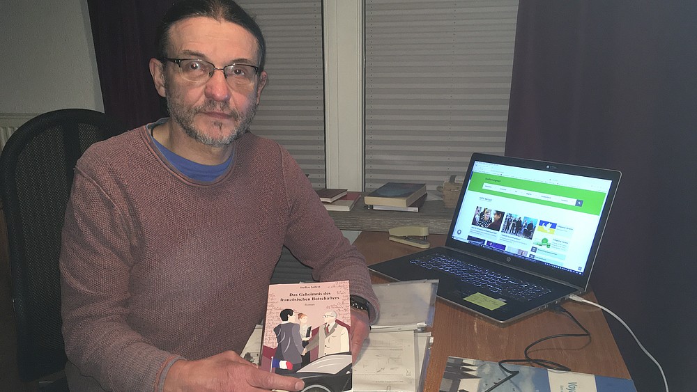 Steffen Seifert hält sein Buch in die Kamera und sitzt am Schreibtisch.