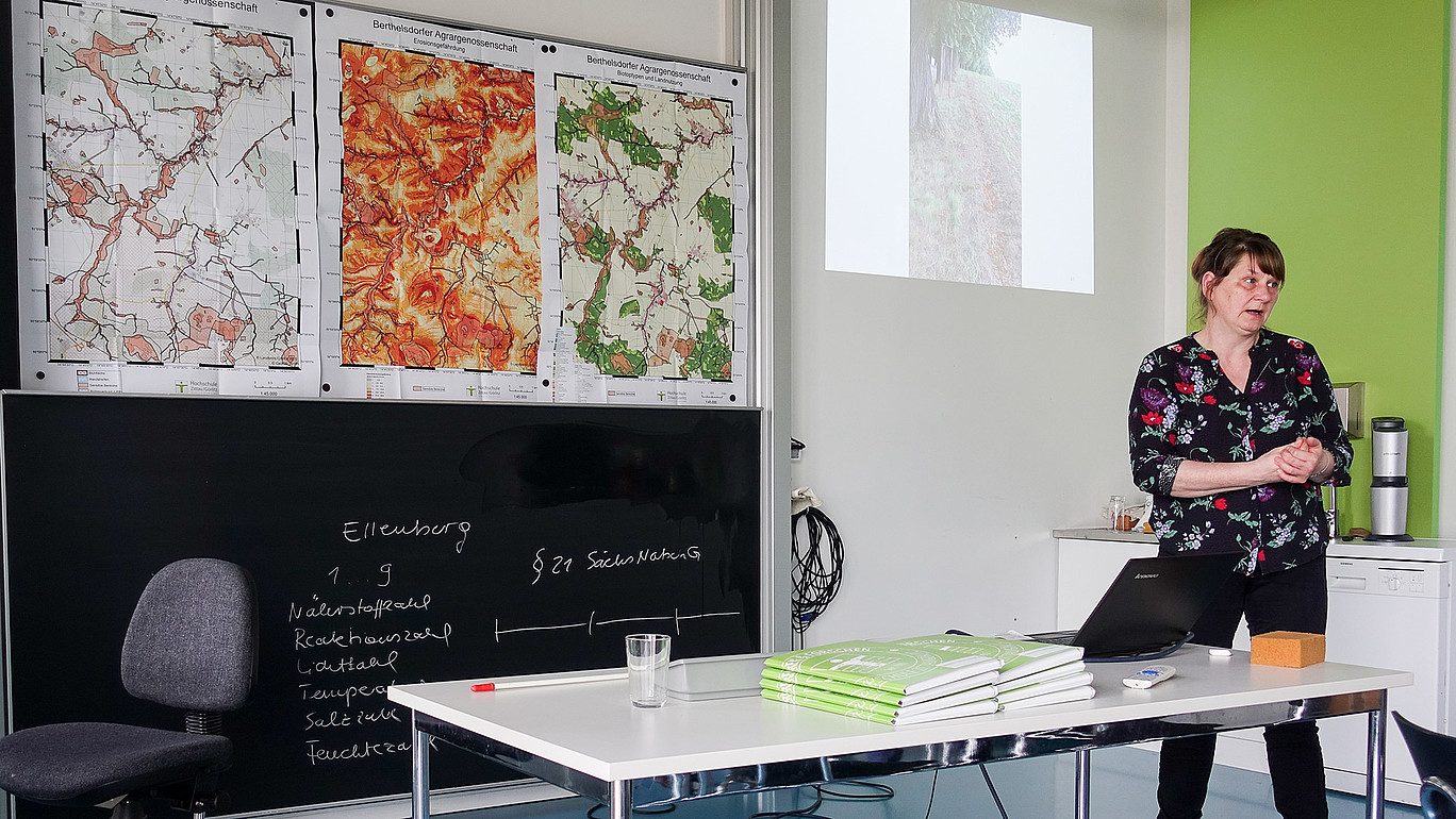 Eine Frau steht vor einer Tafel und spricht.
