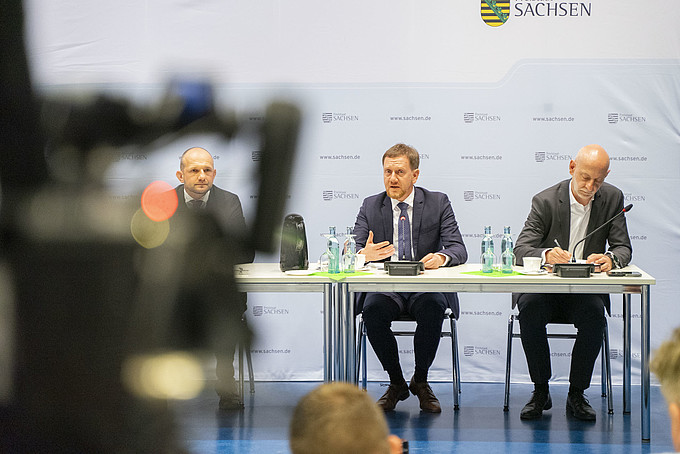 Fokussierte Ansicht von Landrat Meyer, Ministerpräsident Kretschmer und Regierungssprecher Schreiber bei der Pressekonferenz, Kretschmer spricht betont, flankiert von Meyer und Schreiber, mit Mikrofonen und Wasserflaschen auf dem Tisch.