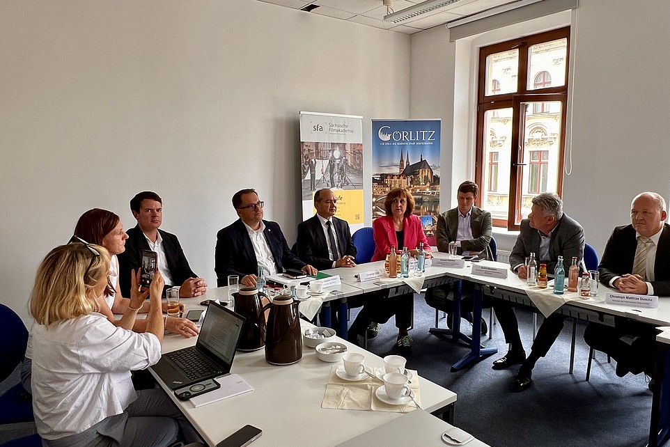 Die Gesellschafterinnen und Gesellschafter sitzen in einem Seminarraum an Tischen und besprechen sich.