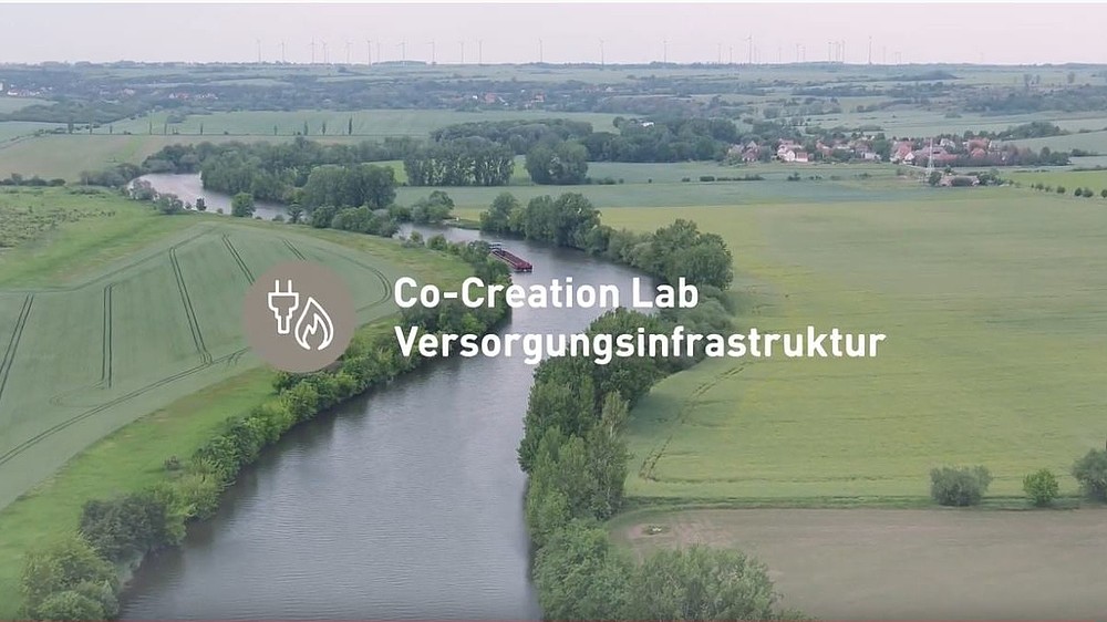 Eine Luftansicht von grünen Feldern, durch die sich die Saale schlängelt mit dem Schriftzug Co-Creation Lab Versorgungsinfrastruktur ist zu sehen.