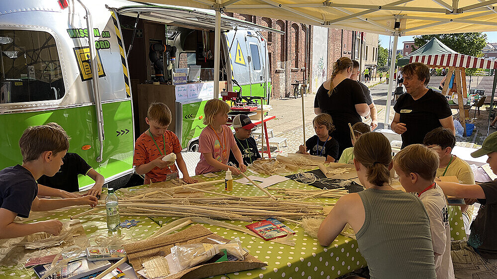An einem großen Tisch sitzen Kinder eine Betreuerin, vor ihnen liegen Naturfasern auf dem Tisch, mit denen sie basteln. Im Hintergrund steht der mobile Lernort-Anhänger der Hochschule.