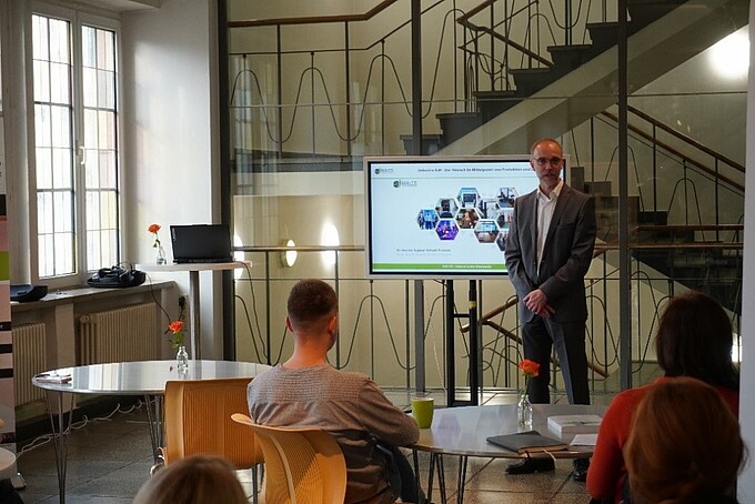 Prof. Wendt spricht neben einem Bildschirm ans Publikum gewandt, das an Rundtischen sitzt.