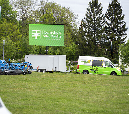 Auf dem Außengelände steht der HSZG-Sprinter mit einem Anhänger, auf dem eine große LED-Leinwand aufgebaut ist, die gerade das HSZG-Logo zeigt.