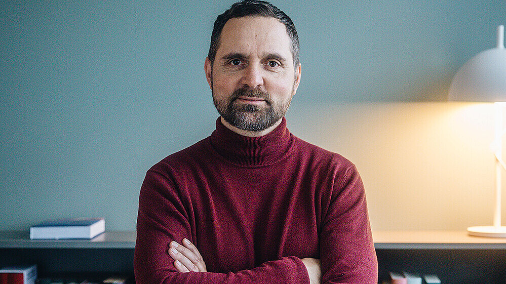 Prof. Sadowski im roten Rollkragenpulli steht lächelnd und mit verschränkten Armen vor einem Bücherregal. Im Hintergrund leuchtet eine Tischlampe.