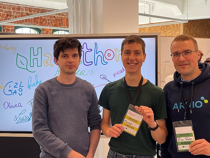 Drei Männer lächeln in die Kamera, im Hintergrund steht ein Bildschirm. Zwei der drei halten ihr Namensschild vor sich.