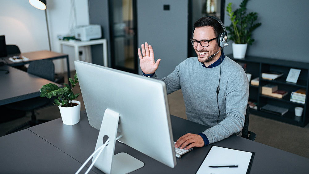 Ein junger Mitarbeiter sitzt im Büro und hebt die Hand lachend zum Gruß vor der Webcam.