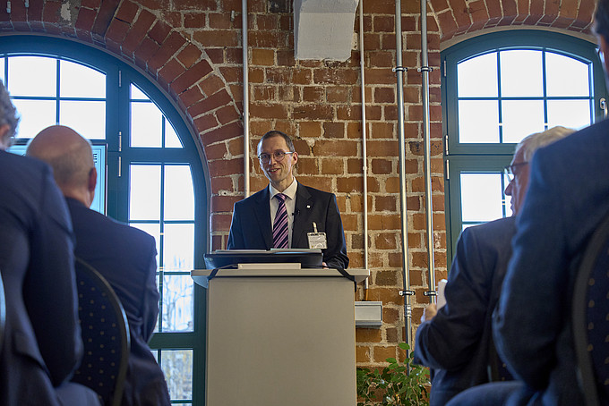 Ein Mann in einem Anzug spricht an einem Rednerpult vor einer Gruppe von Personen in einem Saal mit Backsteinmauern.